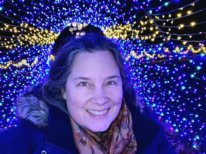 Photograph of Julia Tenney, a white woman with dark hair, in front of a blue & yellow fairy lights display.