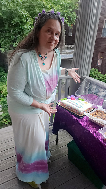 same woman in tie-dyed dress 25 years later, showing off a cake